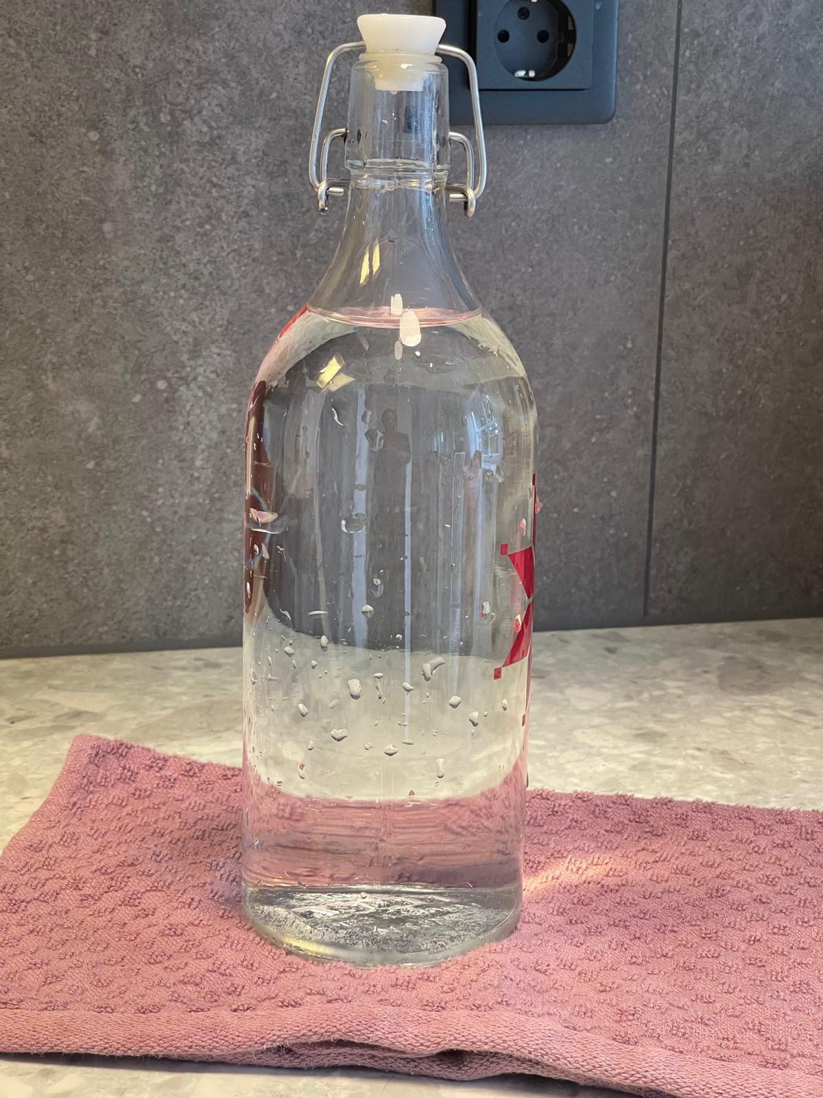 A clear glass water bottle with a narrow neck, sitting on a kitchen counter, ready to be cleaned.